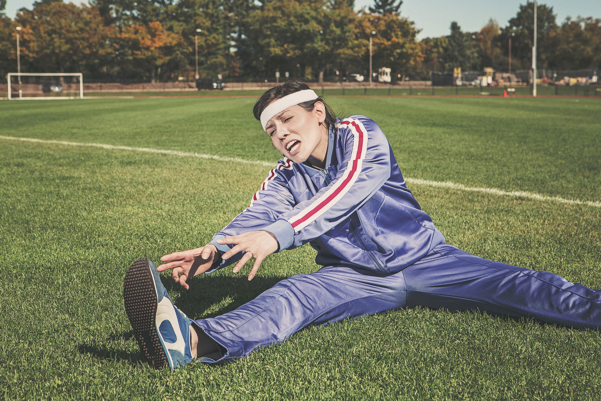 femme en jogging violet, grand écart sur la pelouse d'un terrain, elle a des difficultés.