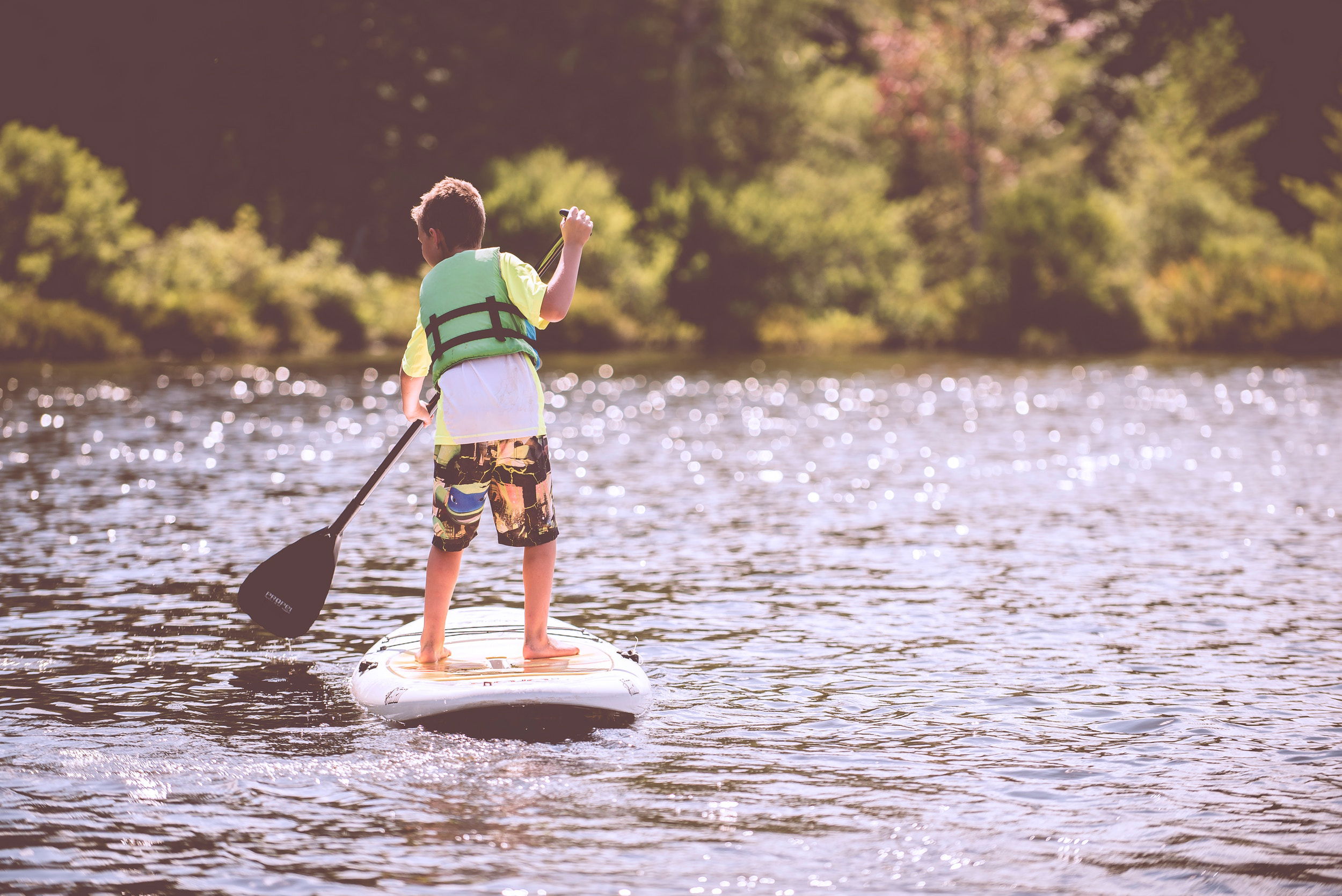A boy is on a paddleboard with a lifejacket on. The background is a beautiful lake with trees.