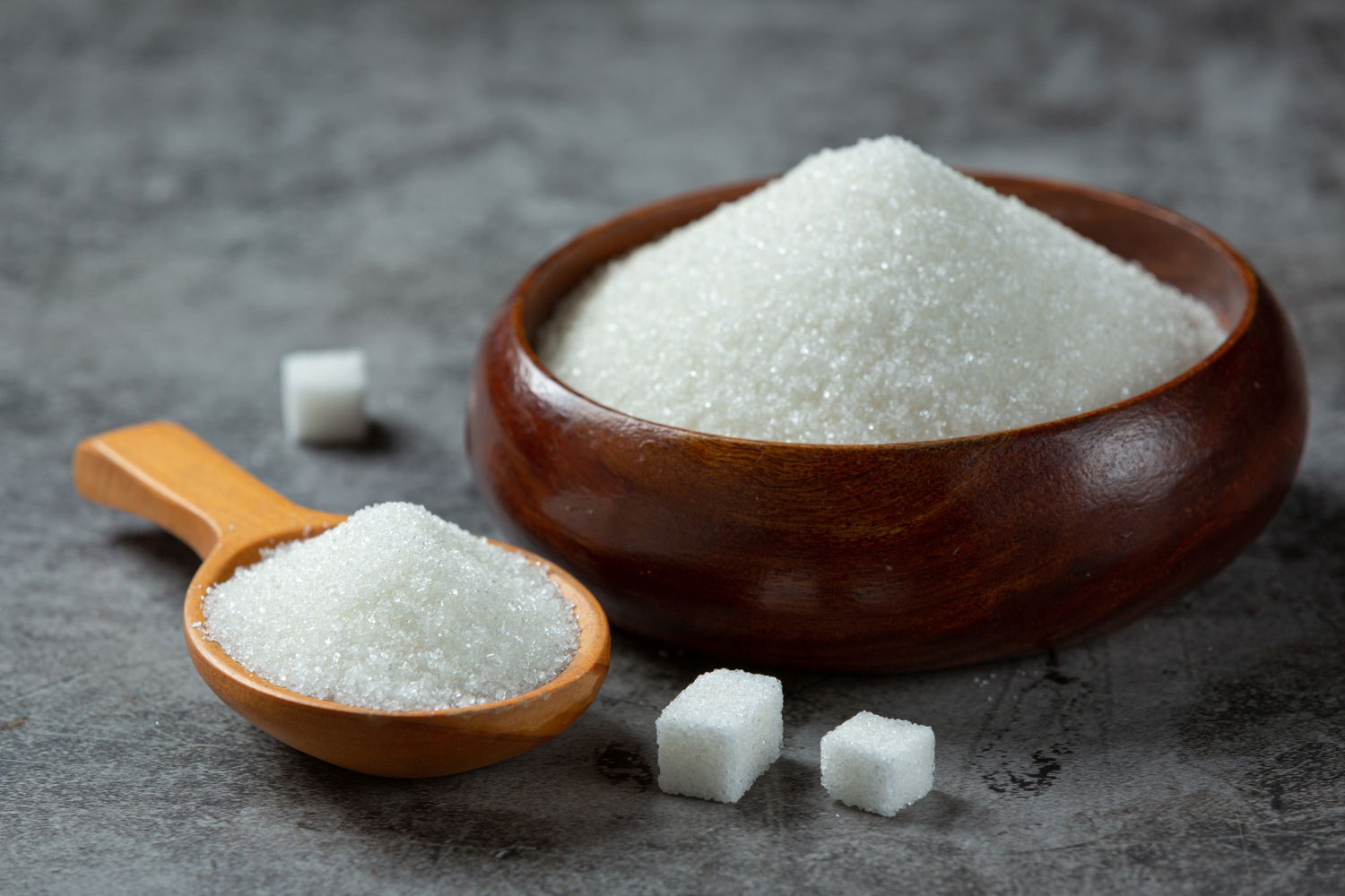 Granulated sugar in a wooden bowl and spoon, with some sugar cubes on the table