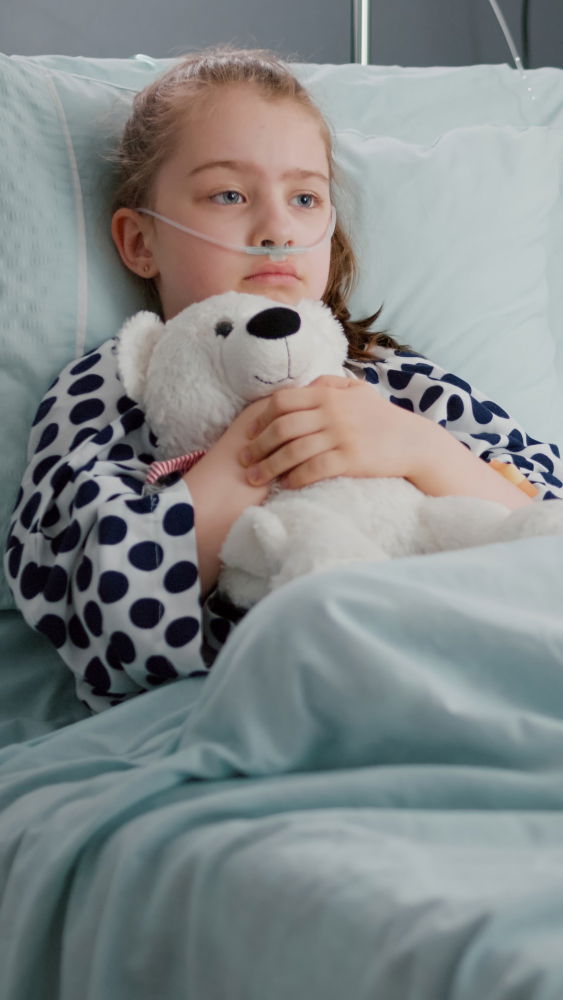  Sick white female toddler lying on a bed, wearing a nasal oxygen tube and holding her doll