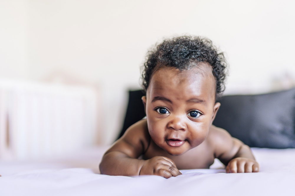 A photograph of a baby lying on their tummy on a soft, light-colored surface. The baby is facing the camera with wide, expressive eyes and a curious, alert expression. The arms are bent at the elbows, supporting their upper body in a tummy-time position.