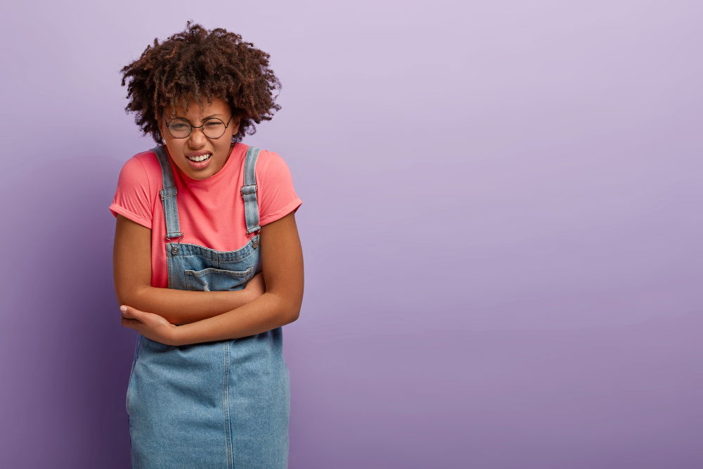 Young woman wearing glasses and a denim overall dress over a pink shirt, standing against a purple background and holding her lower abdomen with a pained expression.