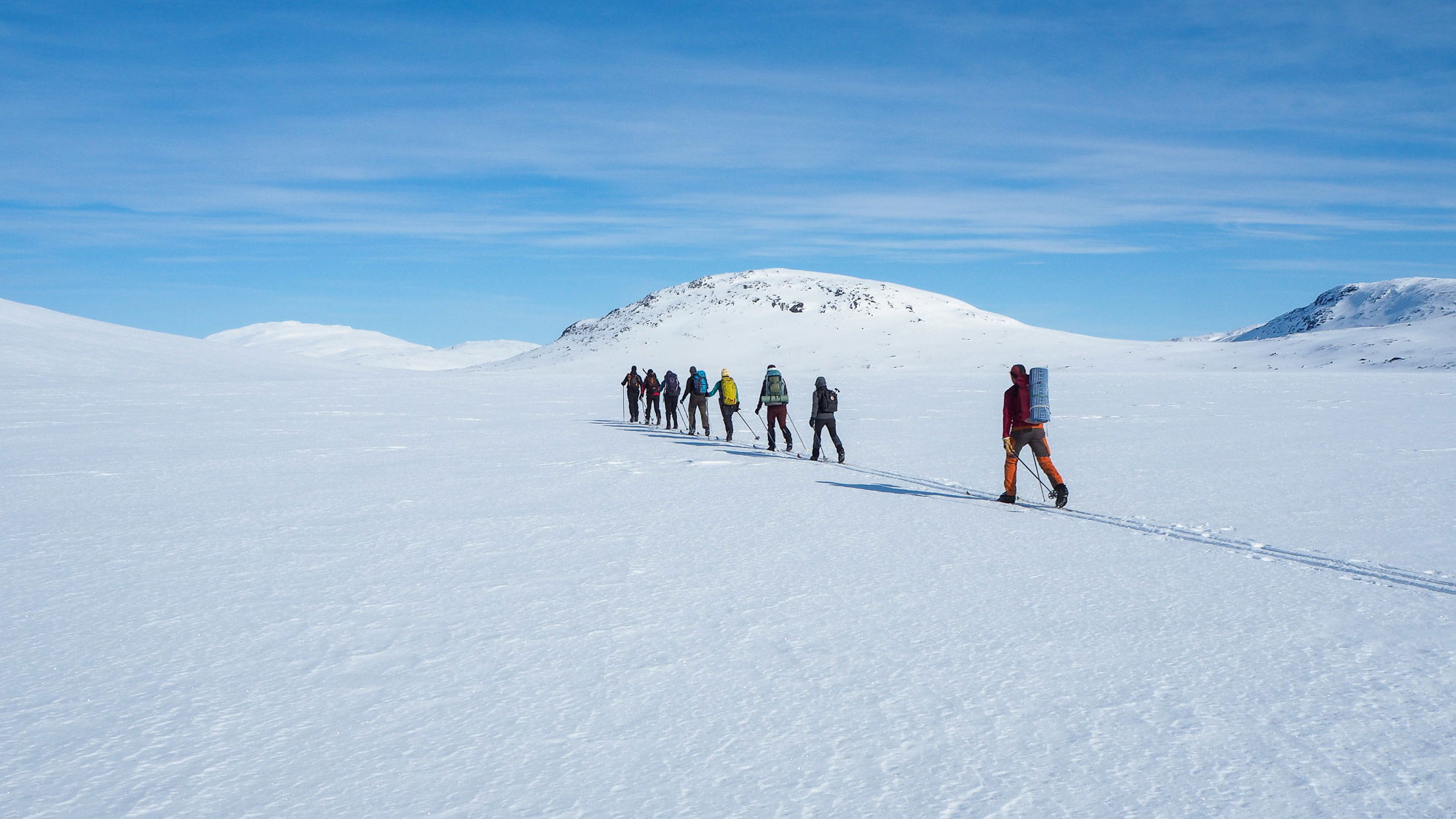 Joukko retkeilijöitä hiihtää jonossa kohti lumisia tuntureita. 