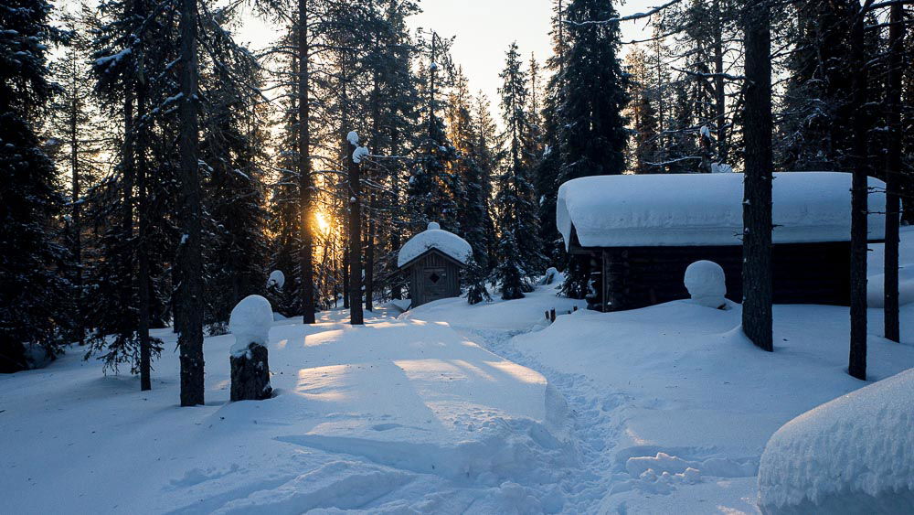 Aikainen aamu Riisitunturin erämaassa. Aurinko on nousemassa puiden takana. Kuvassa lumen peittämä hyyskä ja liiteri. 