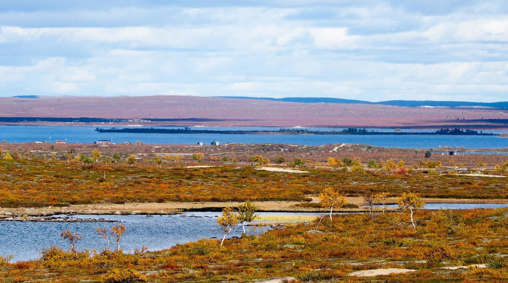 Kuvassa etualalla ruskan värjäämää varvikkoa ja kauempana siintää sininen järvi. Järven rannalla muutama pieni kesämökki.