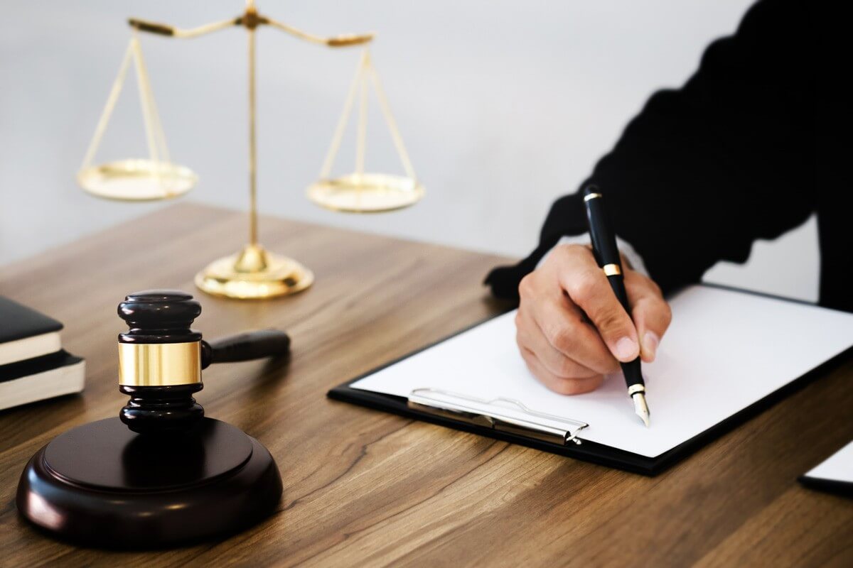 A legal professional writing on a document with a judge's gavel and golden scales of justice on a wooden desk, symbolizing fairness, law, and legal proceedings.