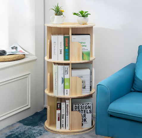 A 3 tier rotating pine book shelf featured in a quaint room with a blue chair and bright sunlight coming in through a window.
