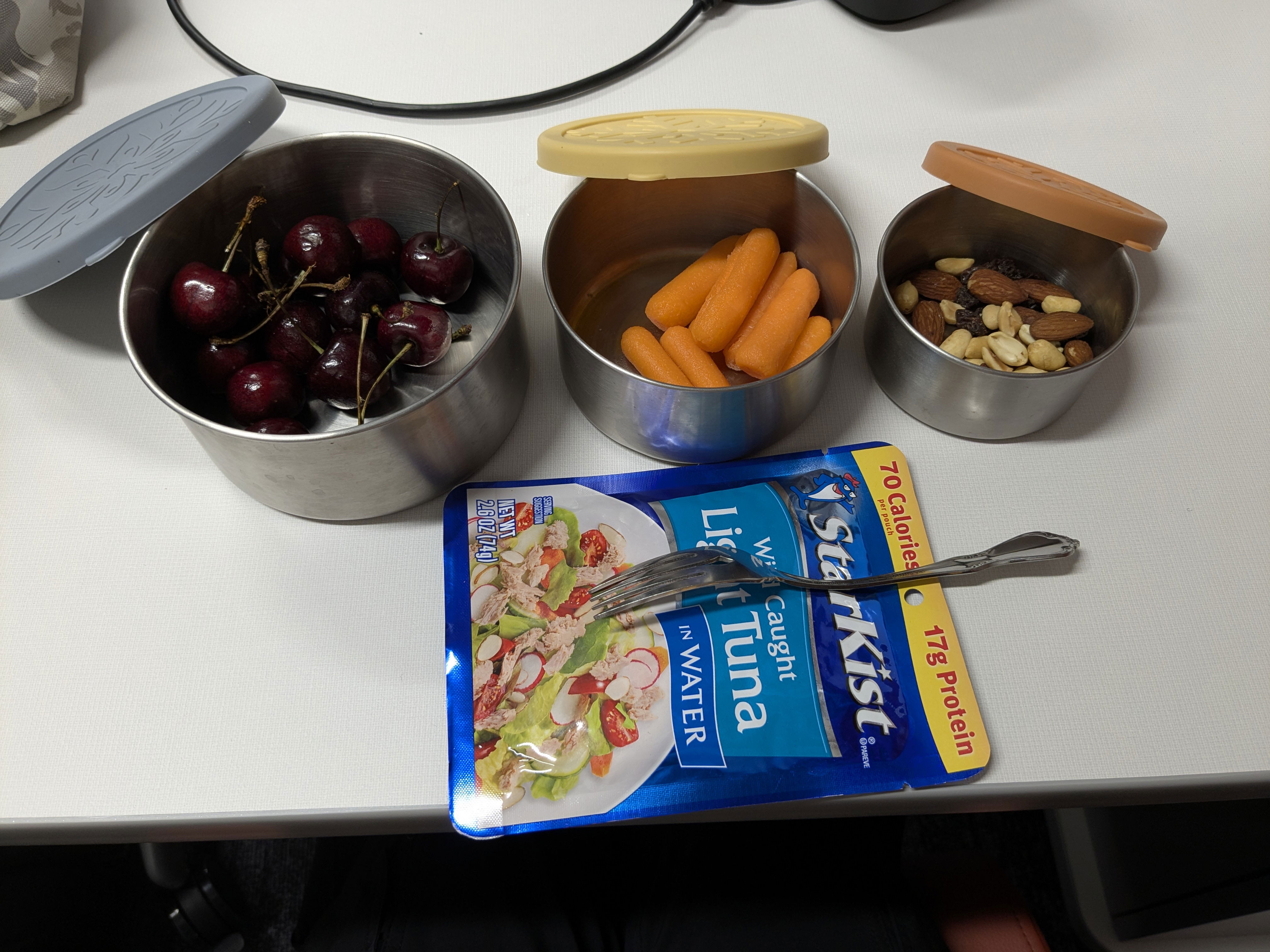 Cherries, carrots and almonds are shown in varying size stainless steel containers along with a package of tuna for a healthy lunch.