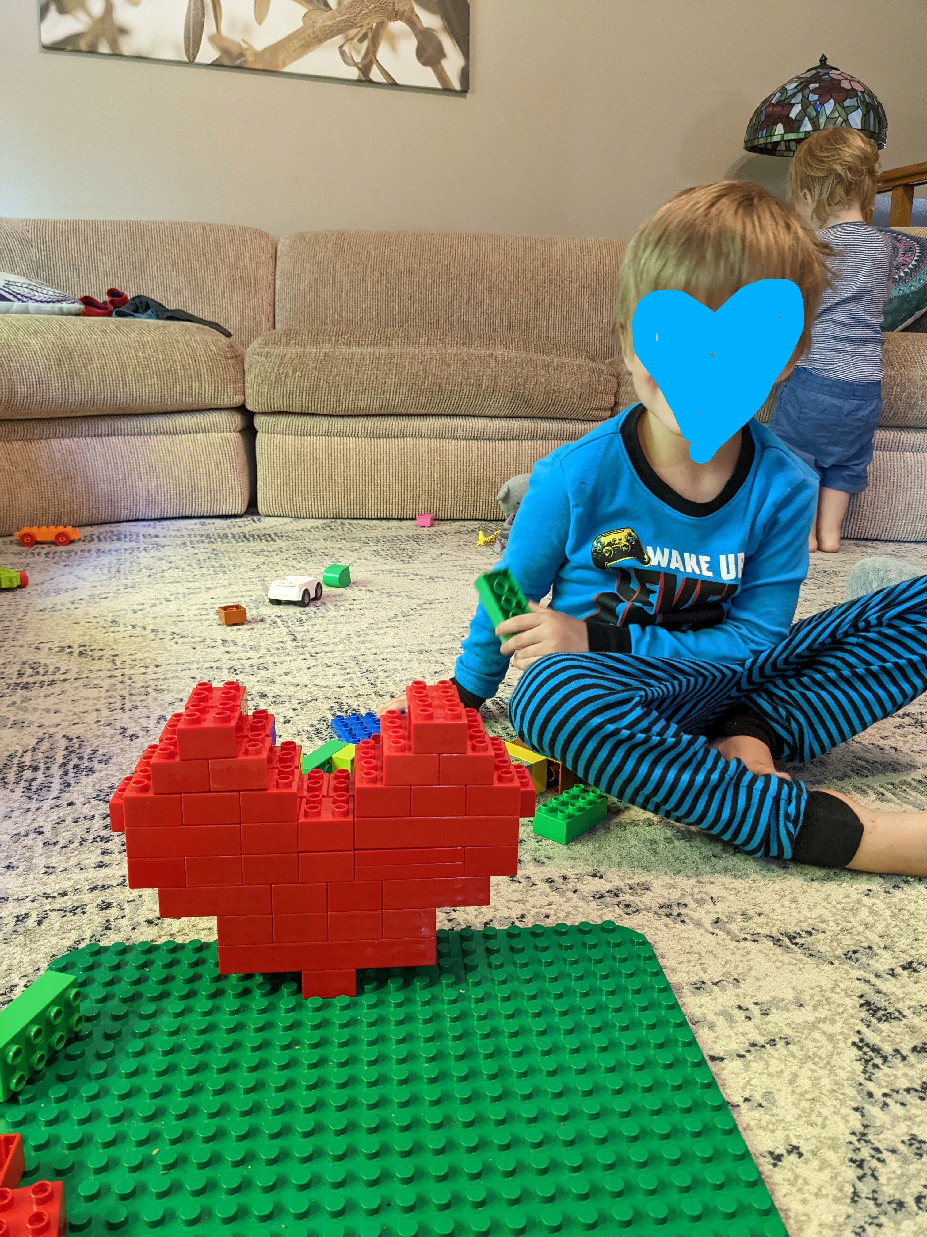 A young boy with his face covered for privacy is sitting next to his Lego duplo build of a big red heart