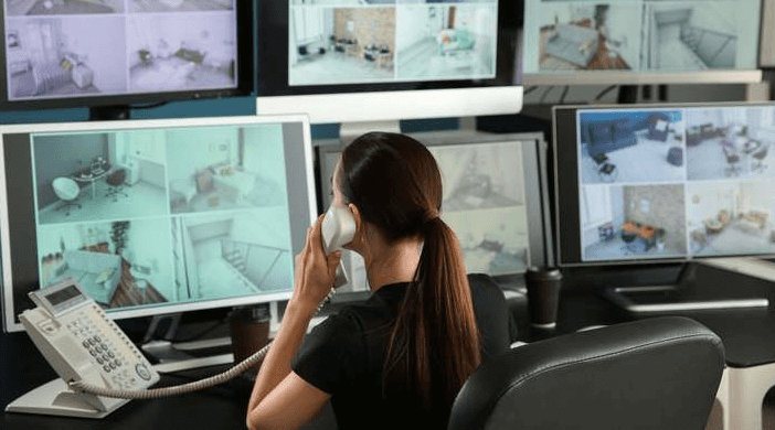 A woman with her back to the viewer is seated at a desk, speaking on a phone while surrounded by multiple computer monitors displaying various CCTV camera feeds, indicating a remote monitoring service.