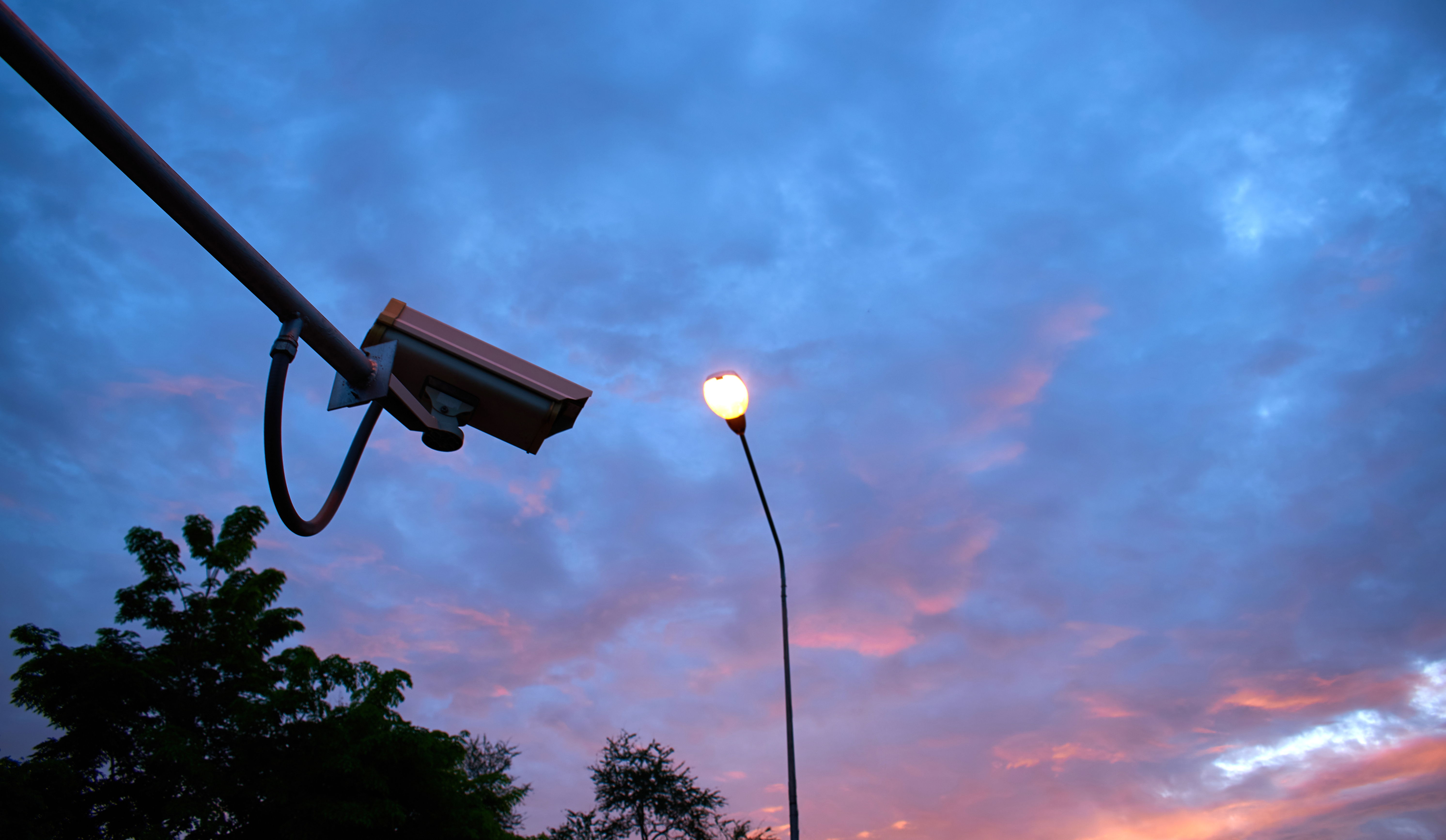 Outdoor bullet-style security camera viewed through rusted metal bars, symbolizing continuous surveillance by a trusted monitoring services provider.