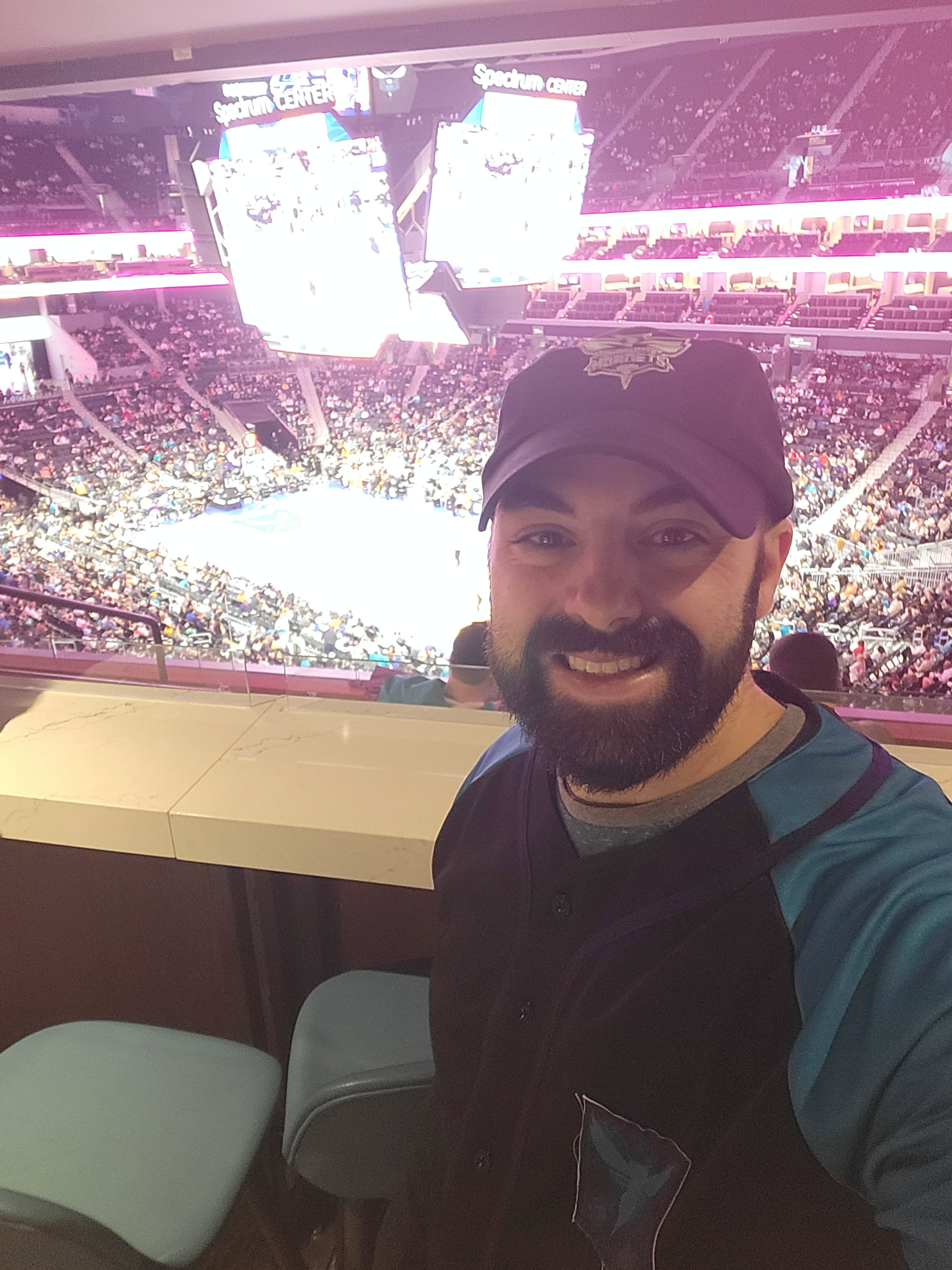 Smiling man takes a selfie inside a packed Spectrum Center while watching a live basketball game.