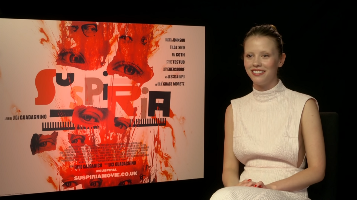 Still of Mia Goth during an interview for Suspiria with a movie poster beside her.