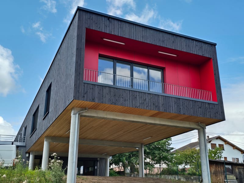 Charpente et murs à ossature bois - Couverture et bardage en zinc à joint debout - Etanchéité sur toiture terrasse avec protection par gravillons et végétalisation - Bardage en bois teinte sombre - Bardage en bois en sous-face - Bardage en Trespa®