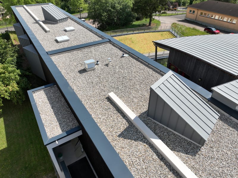 Charpente et murs à ossature bois - Couverture et bardage en zinc à joint debout - Etanchéité sur toiture terrasse avec protection par gravillons et végétalisation - Bardage en bois teinte sombre - Bardage en bois en sous-face - Bardage en Trespa®