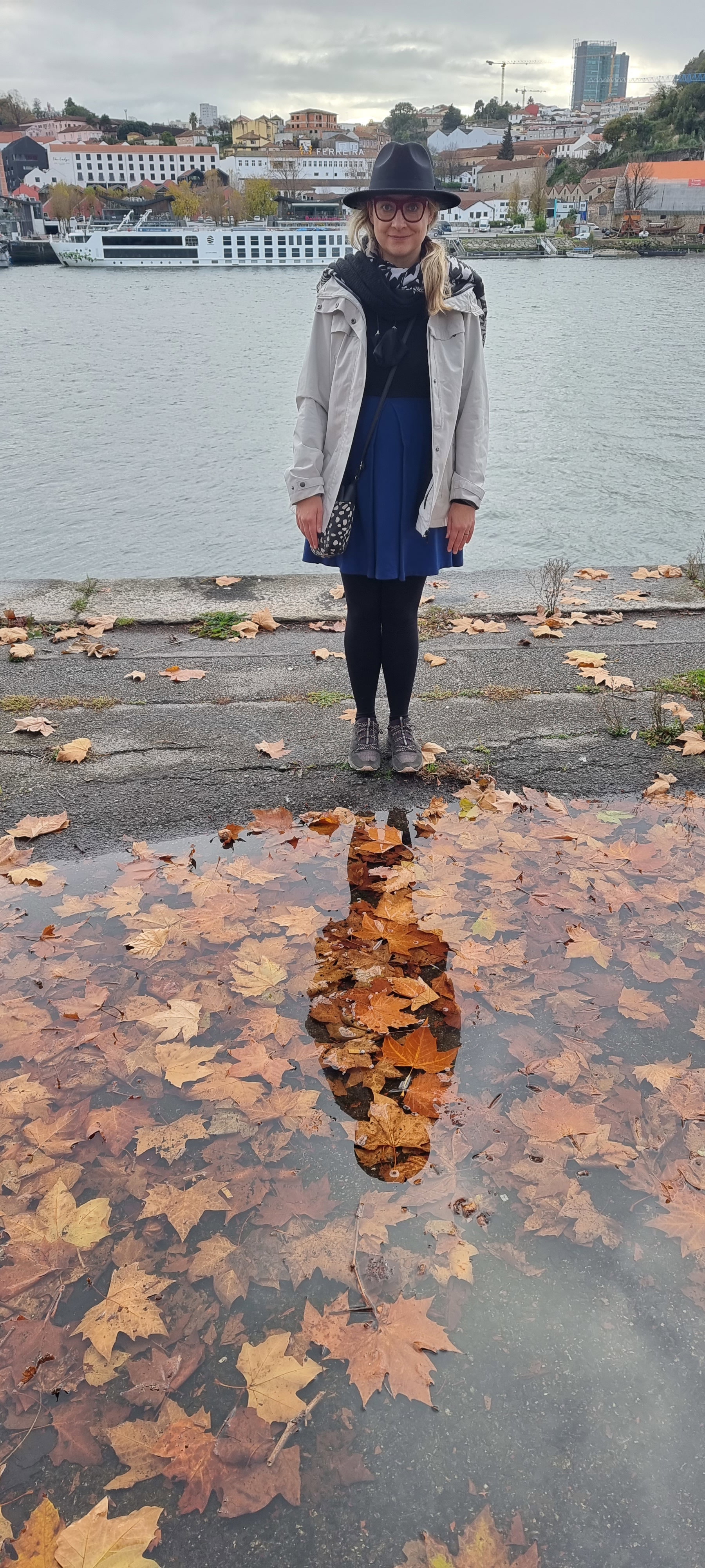 Moira from Portugal Horizon standing by the Douro river in Porto on a grey autumn day, with the Vila Nova de Gaia waterfront and its port wine lodges behind her.