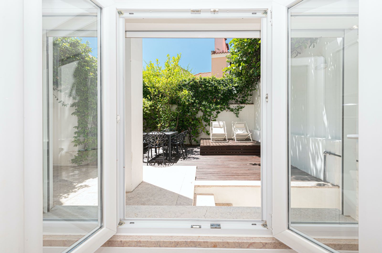 Open glass doors leading from a bright, renovated Príncipe Real studio onto a private garden with loungers and afternoon sun, Lisbon.
