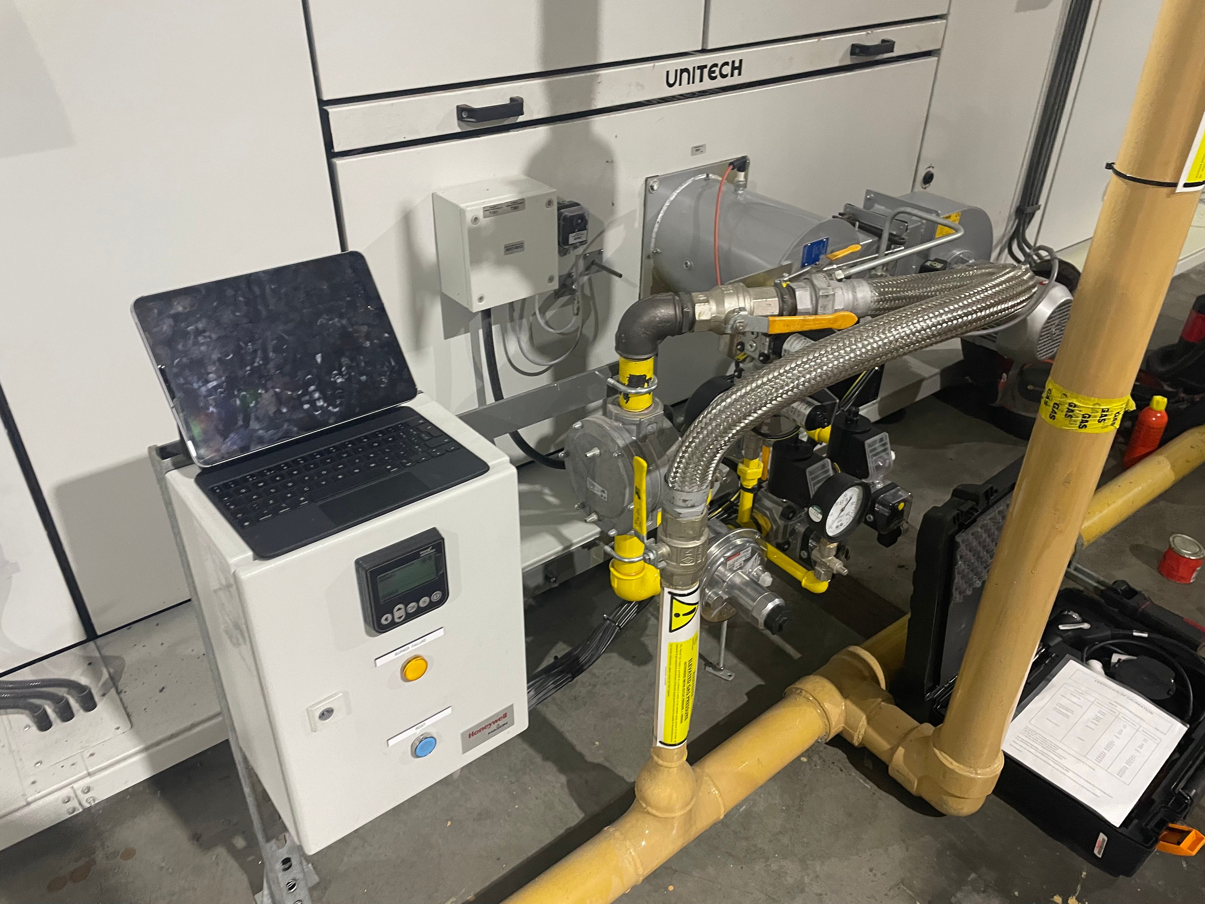 Burner control panel with a tablet on top, beside the burner gas train on a Unitech unit, with a test kit case on the floor