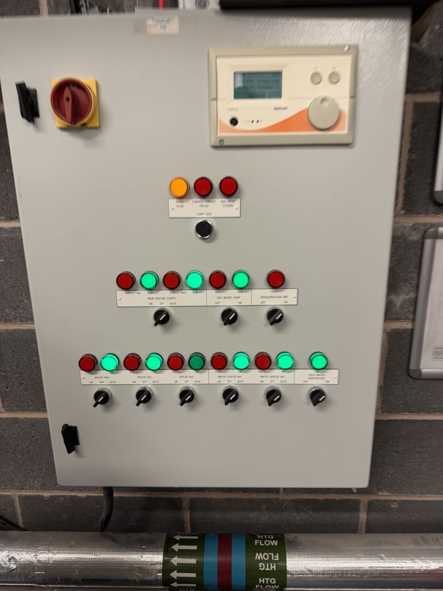 Boiler plant control panel showing boiler, heating pump and hot water pump status lights and selector switches, above insulated heating flow pipework