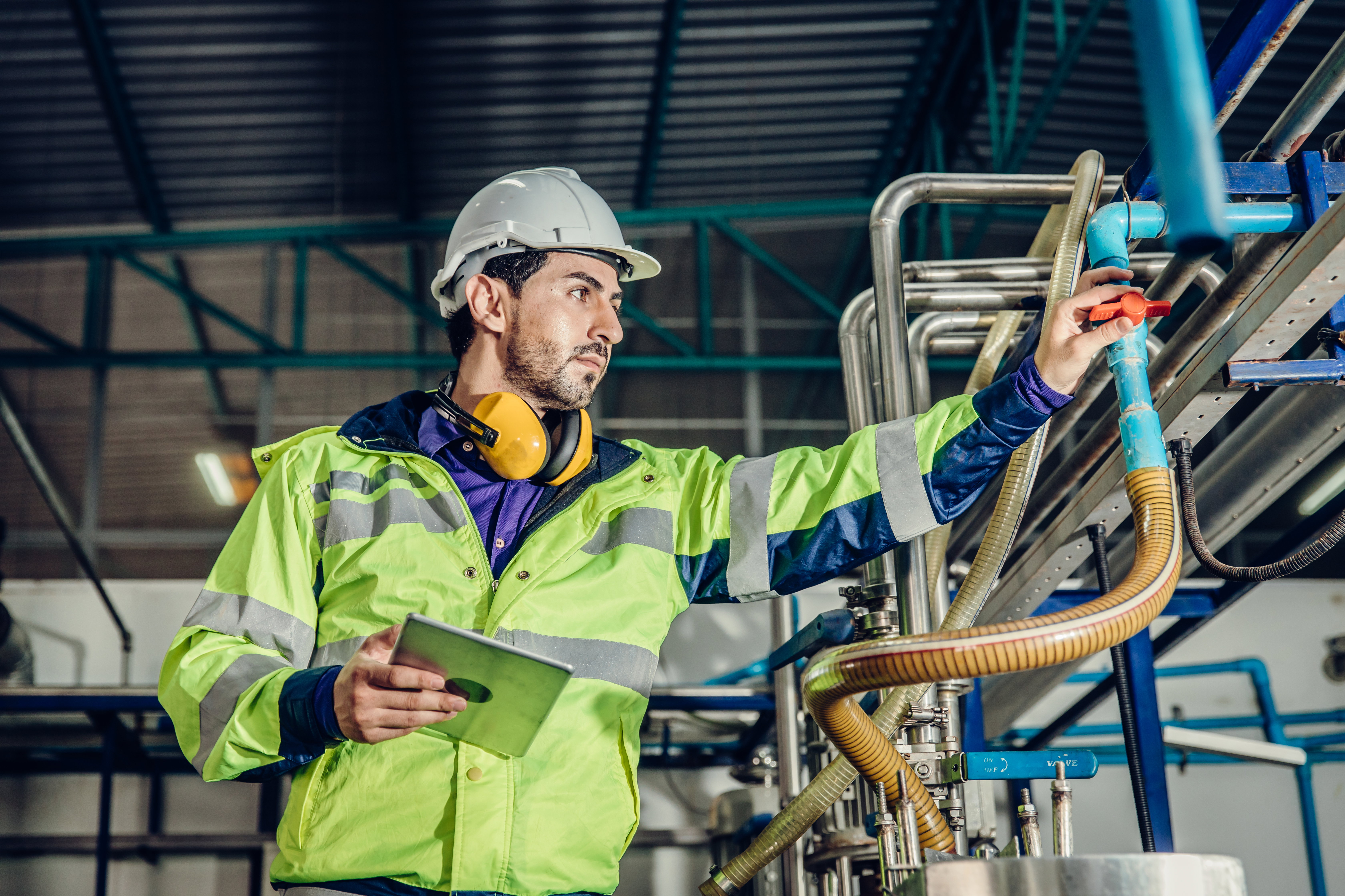 Engineer in a hi-vis jacket and white hard hat adjusting a valve on factory pipework while holding a tablet