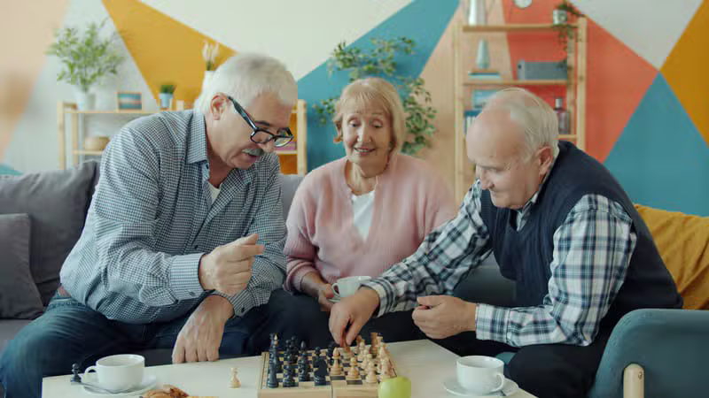 Three older adults playing a friendly game of chess together over coffee in a bright community room