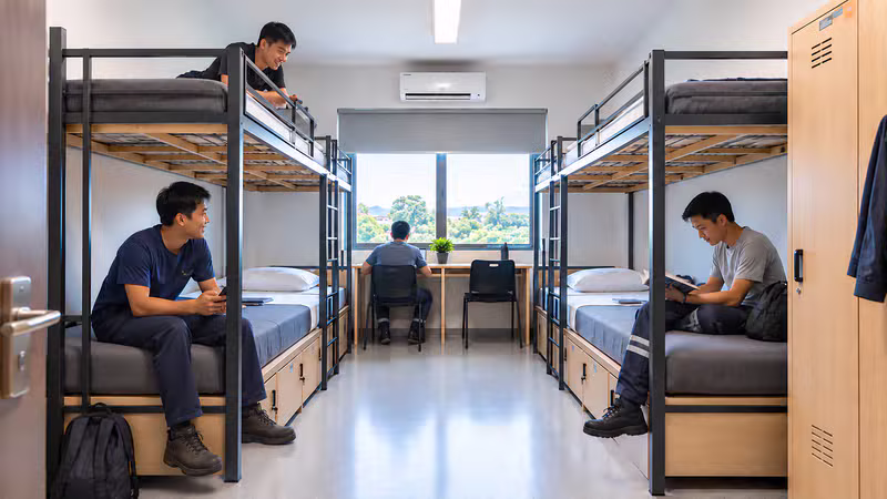 Workers seated on the lower bunks of a dormitory room with metal bunk beds and wooden lockers