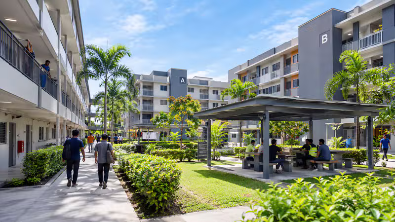 Workers walking along a landscaped courtyard path between multi-storey accommodation blocks with palm trees and a shaded seating shelter