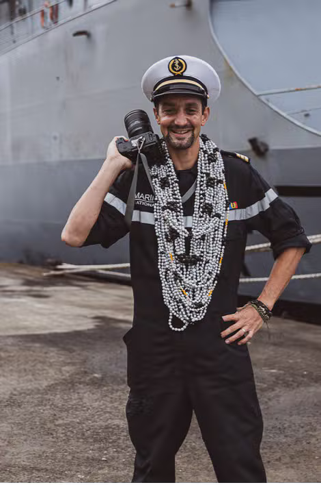 Julien Meillon, photographe en Guadeloupe, en tenue de marin sur le pont d'un navire, appareil photo à l'épaule