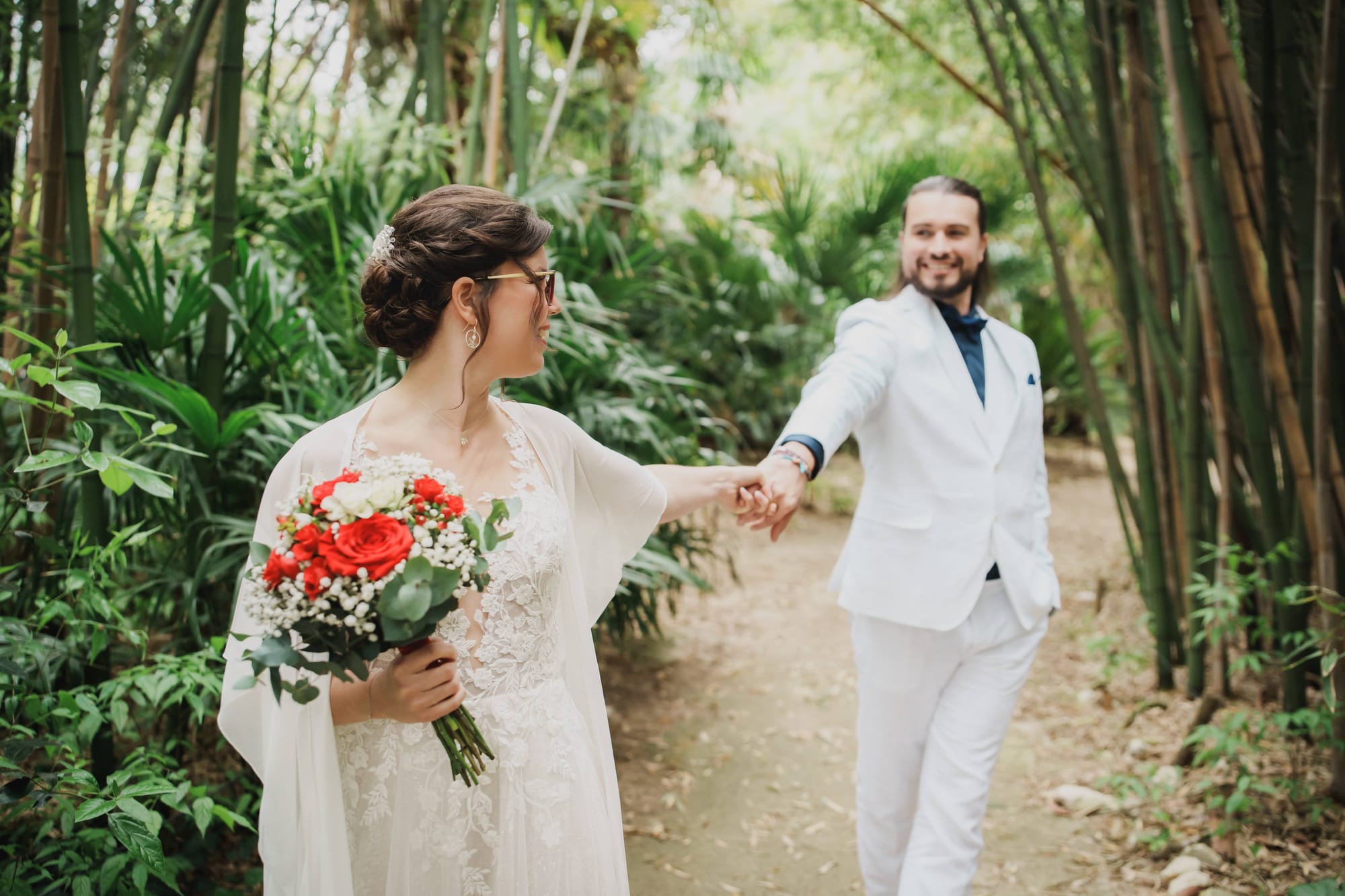 Chignon de mariée tressé élégant, bijou perlé. Robe blanche en dentelle florale avec manches fluides. Bouquet rouge, blanc et vert aux roses éclatantes.