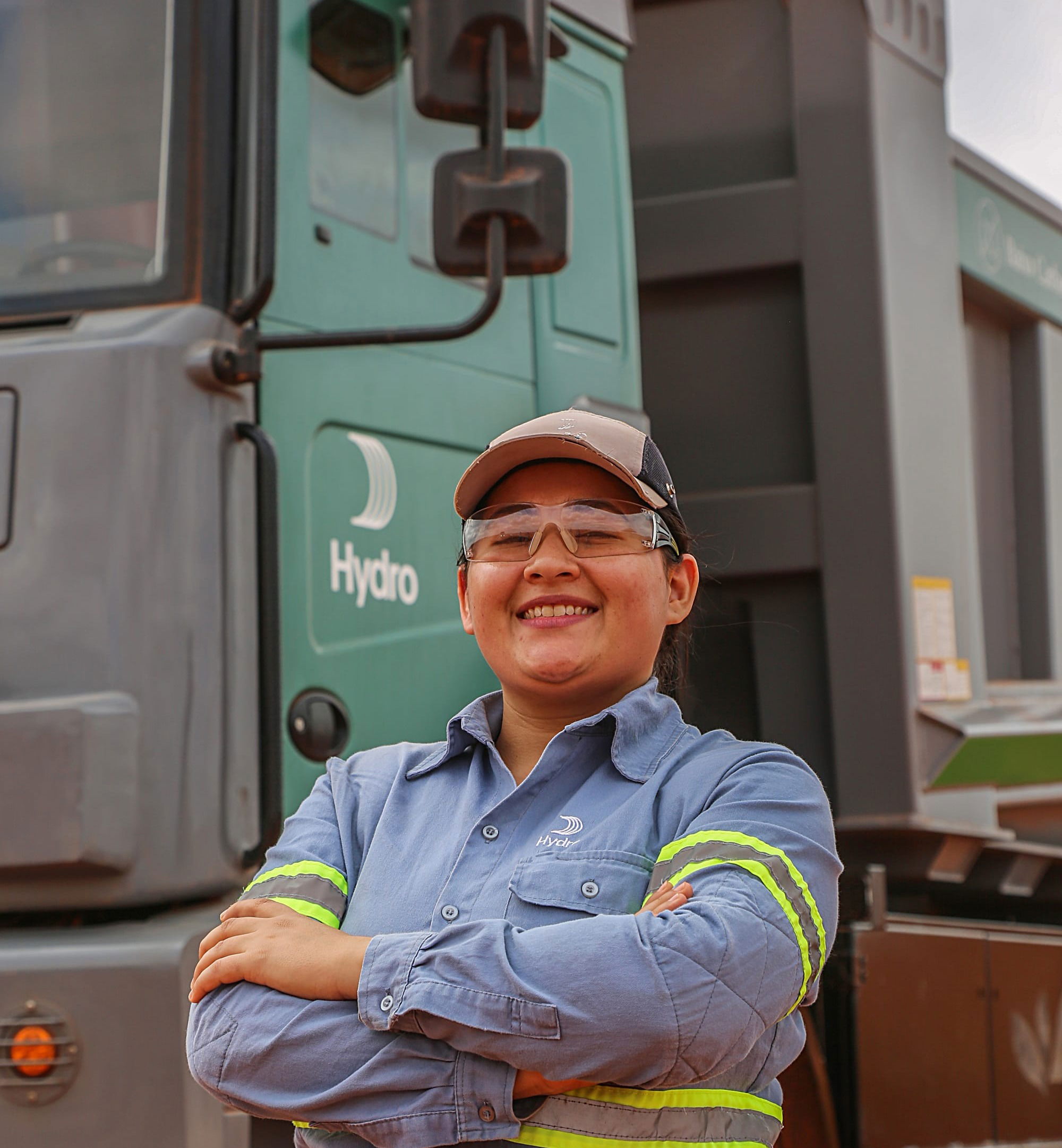 Mullher no centro da foto, sorrindo, trajando equipamentos de proteção individual. Ao fundo um caminhão elétrico com a identidade visual escrito Hydro