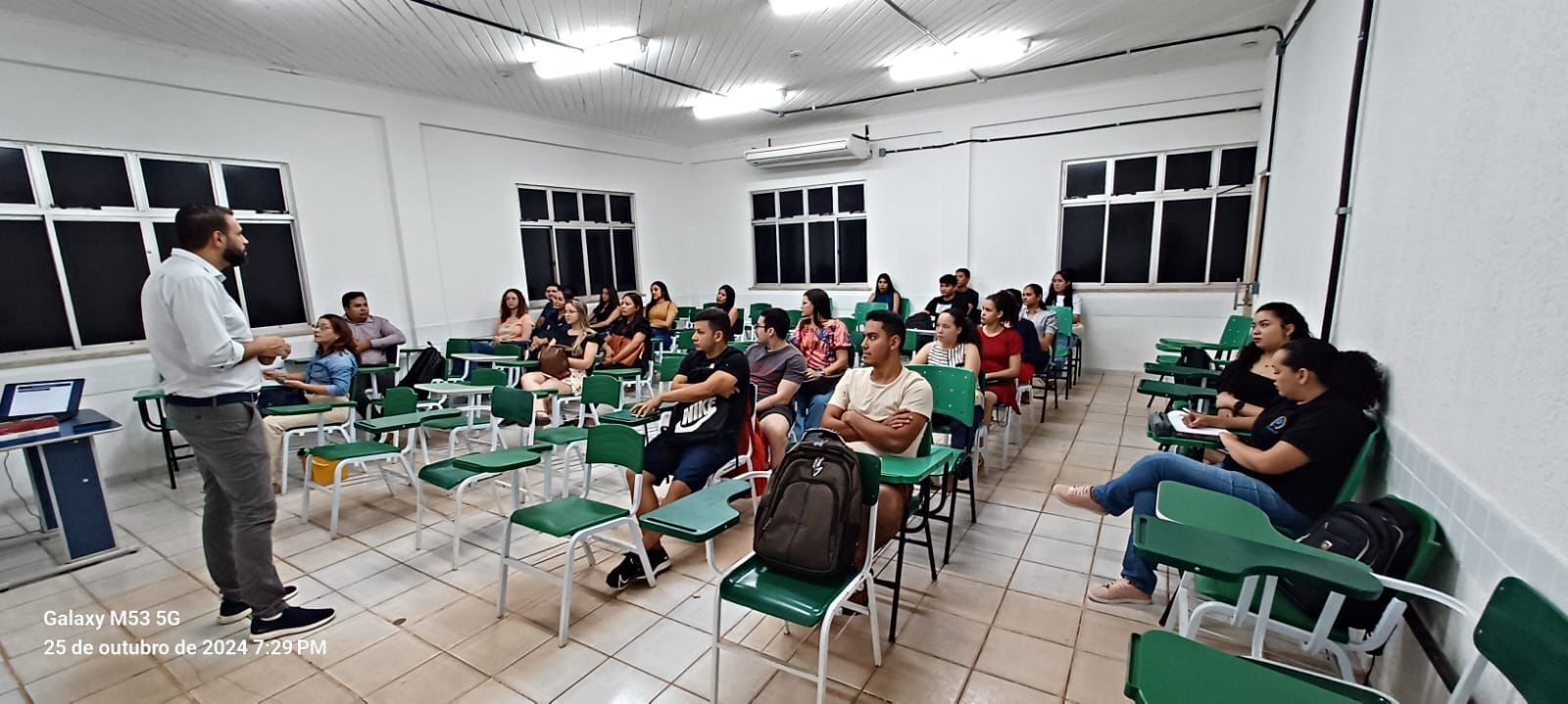 Pessoas em uma sala de aula, sentadas em cadeiras da cor verde participando de uma palestra, onde no centro da foto existe um homem de pé