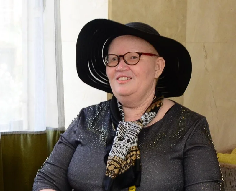 Jeriter Mutinda wear a floral dress and a wide breamed hat. She is seated in a chair.