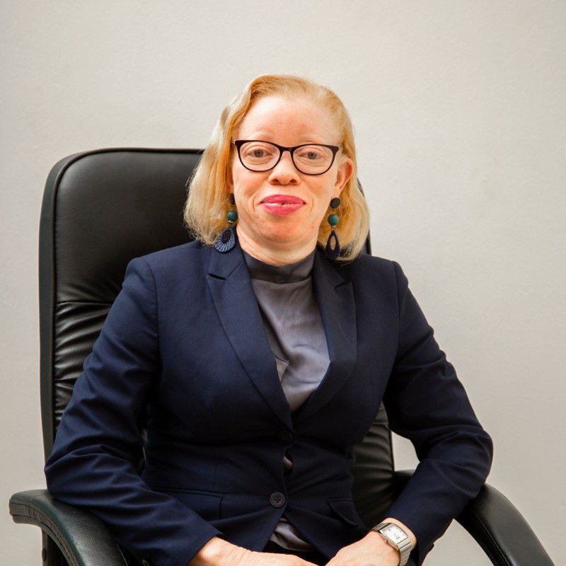 Olive Namutebi sitting on a black chair, dressed in a black blazer and a grey blouse