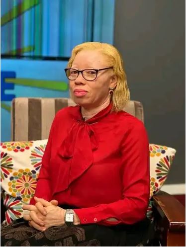 Olive Namutebi dressed in a red blouse seated on a couch with flowery and colourful pillows behind her