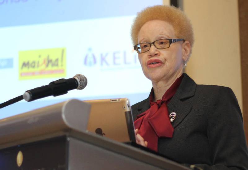 Justice Mumbi Ngugi in a black coat and a red scarf standing on a podium in front of a microphone