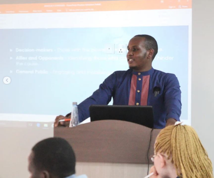 A picture of Boniface Mirikiwa, our project officer training on Advocacy. He is standing wearing a navy blue shirt looking at the participants.