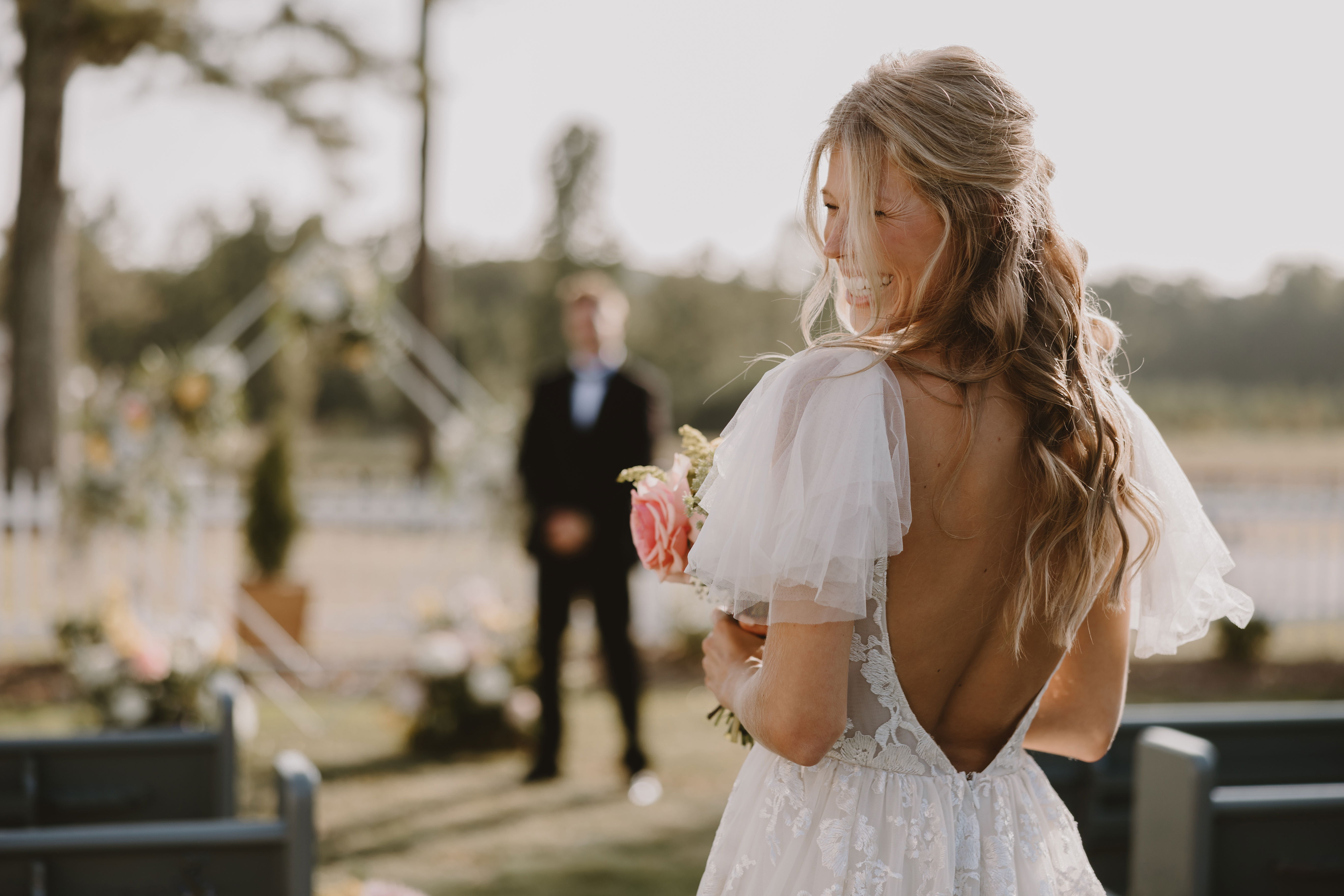 bride entering courtyard for outdoor wedding ceremony with geometric arch, church pews, flowers overlooking Turkey Creek Mountain with views of horses grazing in a pasture near a stable and carriage house and overlooking beautiful countryside