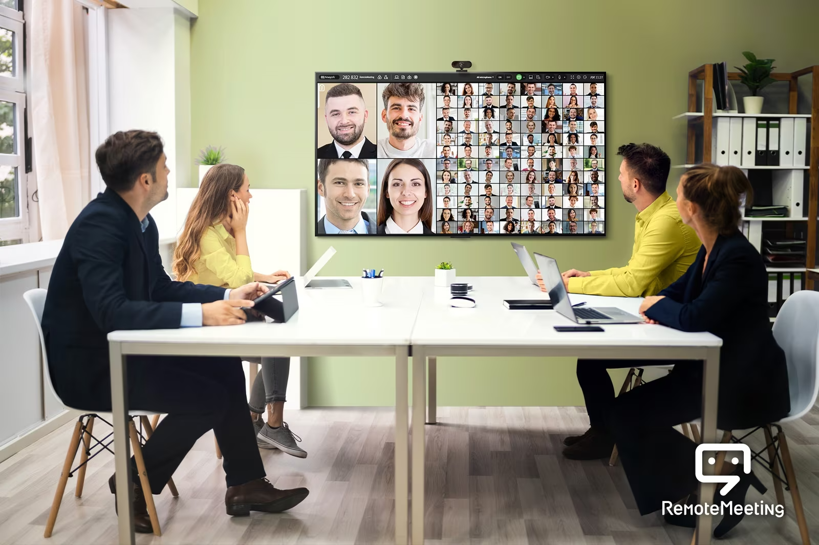 Quatre personnes sont assises dans la salle de conférence sont en train de regarder la télévision et de suivre une téléconférence. L’écran de télévision montre les visages des personnes qui ont participé à la réunion.