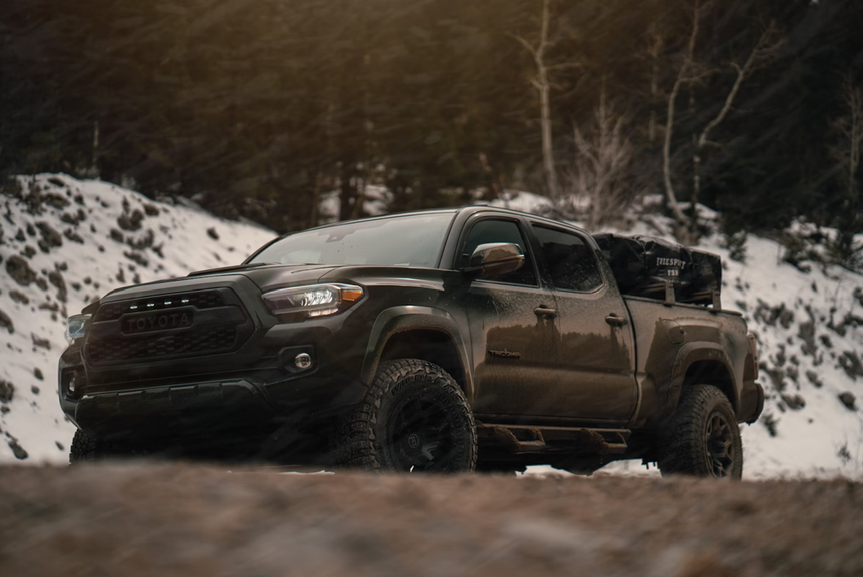 a black truck driving on a dirt road with snow on the side