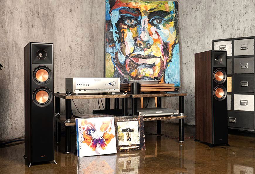 תמונה של זוג רמקולים Klipsch RP-5000F II עומדים משניי צידי שידת מערכת סטריאו עם תקליטים מונחים על הרצפה צמוד לשידה