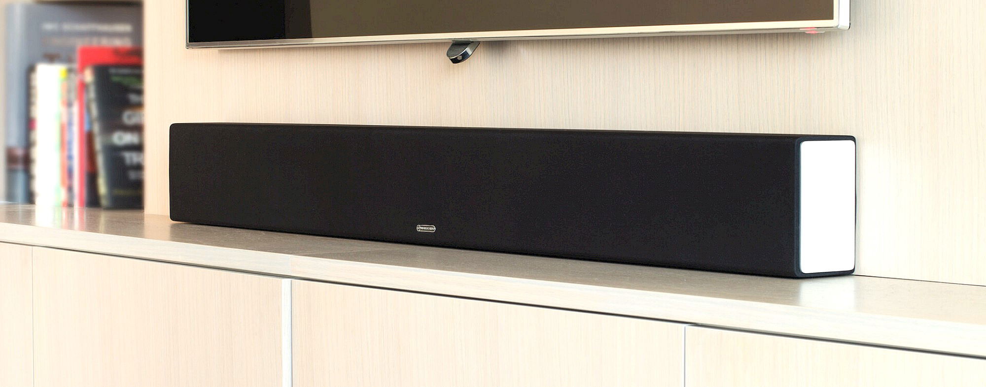 תמונת אווירה של ה Monitor Audio SB-3 סאונדבר מונח על שידה לבנה ומעליו תלויה טלויזיה