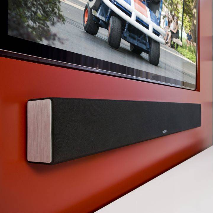 תמונת אווירה של ה Monitor Audio SB-3 סאונדבר תלוי על שידה לבנה ומעליו תלויה טלויזיה וקיר רקע בצבע אדום