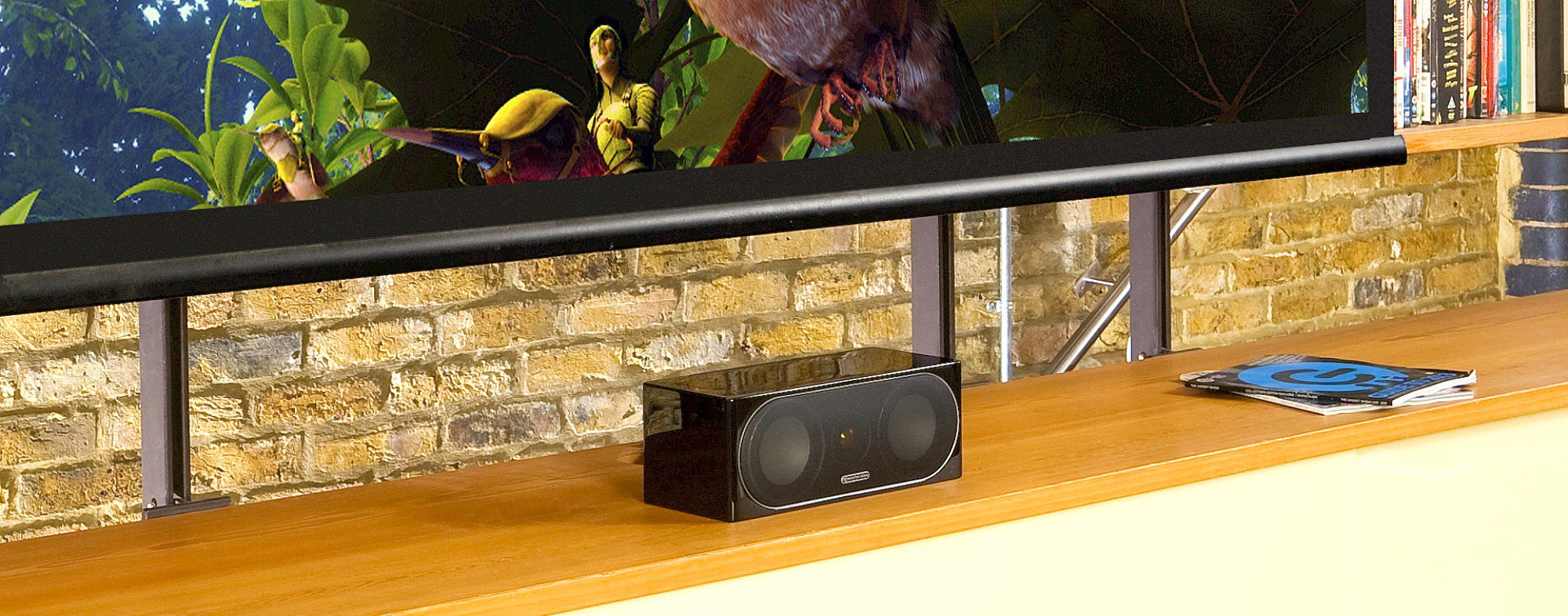 תמונה של הרמקול סנטר מוניטור אודיו Monitor Audio Radius 200 3G בגימור שחור מבריק מונח על שידת עץ בסלון מתחת לטלויזיה עם רקע של קיר לבנים