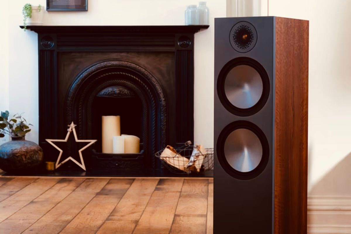 תמונת אווירה של רמקול Monitor Audio Bronze 500 6G בגימור עץ מונח על רצפת עץ מקדימה ומאחרוה ברקע אח