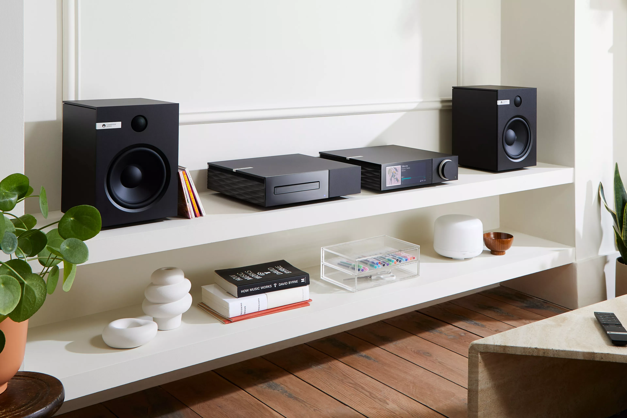 מערכת הכוללת מגבר רסיבר סטרימר היי אנד קמברידג' אודיו Cambridge Audio EVO 150, נגן דיסקים תואם וזוג רמקולים מדפיים קמברידג' אודיו תואמים יושבים על שידת יעץ לבנה בסלון מודרני