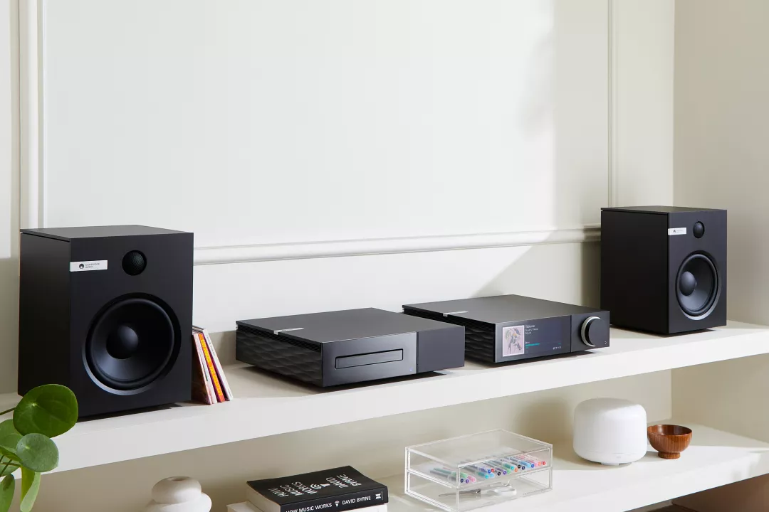 מערכת הכוללת מגבר רסיבר סטרימר היי אנד קמברידג' אודיו Cambridge Audio EVO 75, נגן דיסקים תואם וזוג רמקולים מדפיים קמברידג' אודיו תואמים יושבים על שידת יעץ לבנה בסלון מודרני