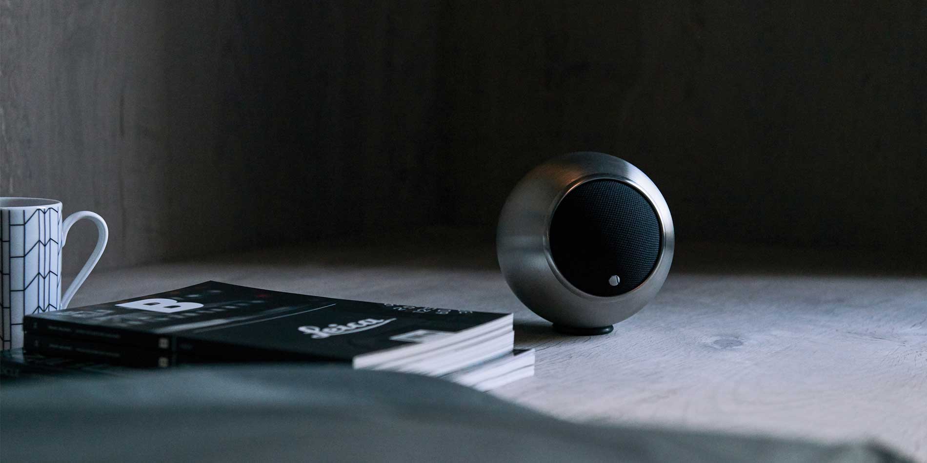 תמונה של רמקול בודד מעותצב גאלו Gallo Acoustics A’Diva בגימור נירוסטה מונח על יד ספר וספל קפה