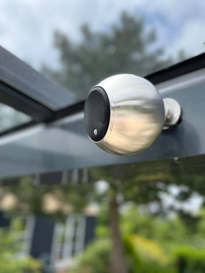 רמקול גאלו Gallo Acoustics Adiva Habitat בצבע נירוסטה תלוי בתוך פרגולה בחוץ