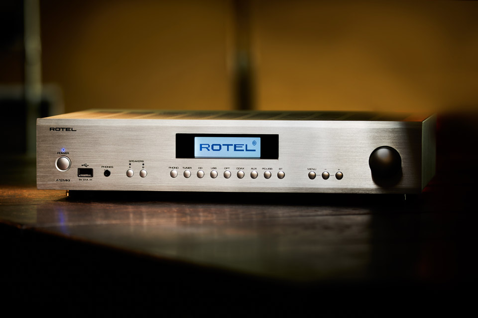 מגבר סטריאו משולב Rotel A12MkII בגימור כסף על משטח עץ, מציג את תצוגת ה-LCD הגרפית, כניסת ה-USB הקדמית ומערך בוררי הכניסות