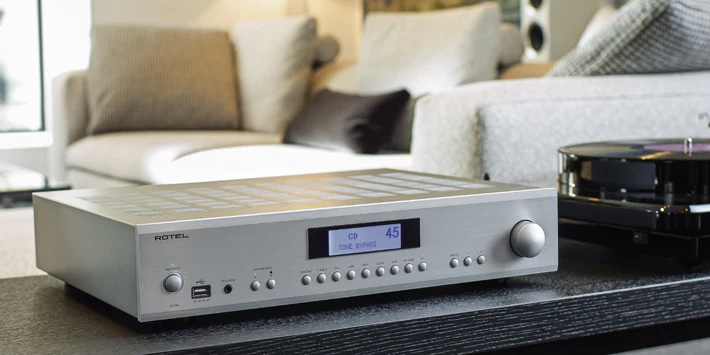 מגבר סטריאו משולב Rotel A14MkII בגימור כסף על שידה מעוצבת בסלון מודרני, לצד פטיפון ותצוגת LCD מפורטת